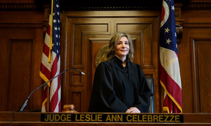 Judge Leslie Ann Celebrezze, a White woman wearing a judge’s robe, stands between two American flags. A plaque on the desk in front of her reads: “Judge Leslie Ann Celebrezze.” 