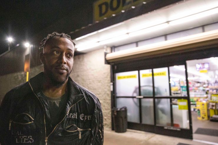 Reginald Dean, a Black man, stands in front of a Dollar General store at night.  