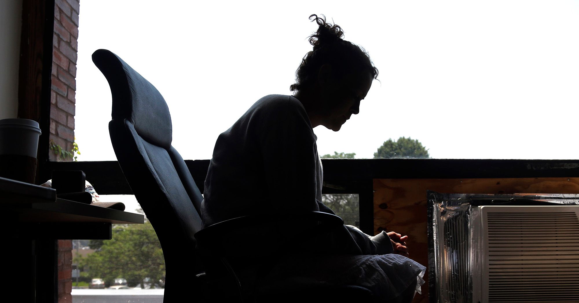 A woman sits in silhouette in front of a large glass window. She's inside an office, sitting on a chair and hunched over with her hands folded on her lap.