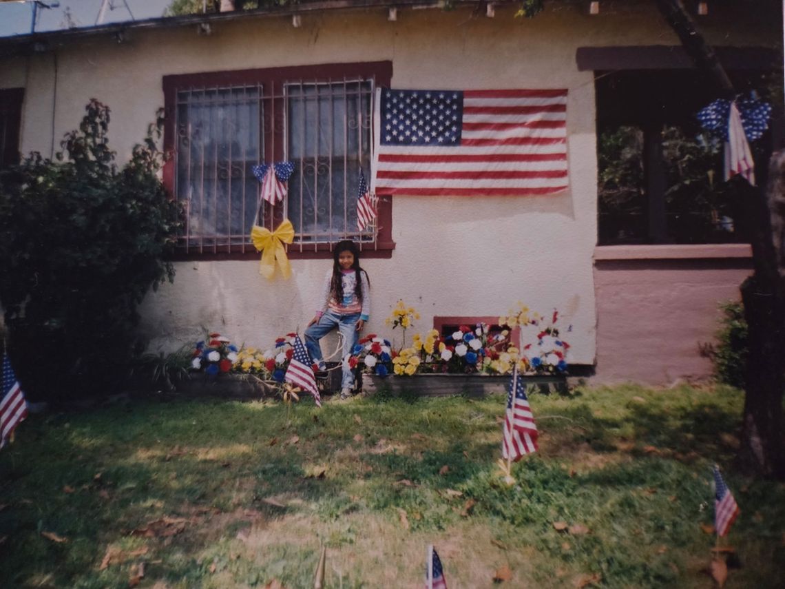 A little girl with long dark hair stands against the outside wall of a one-story house, just beneath a window, an American flag, and a makeshift memorial of yellow ribbons, flag ribbons and flags stuck into the grass. 