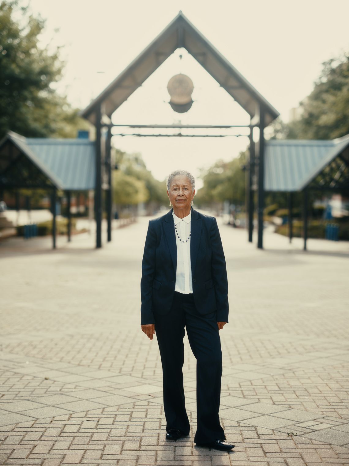 A photo shows an older Black woman with grey hair looking into the camera for a portrait. The woman is wearing a blue jacket and a white blouse, and is standing in the middle of an outdoor plaza.