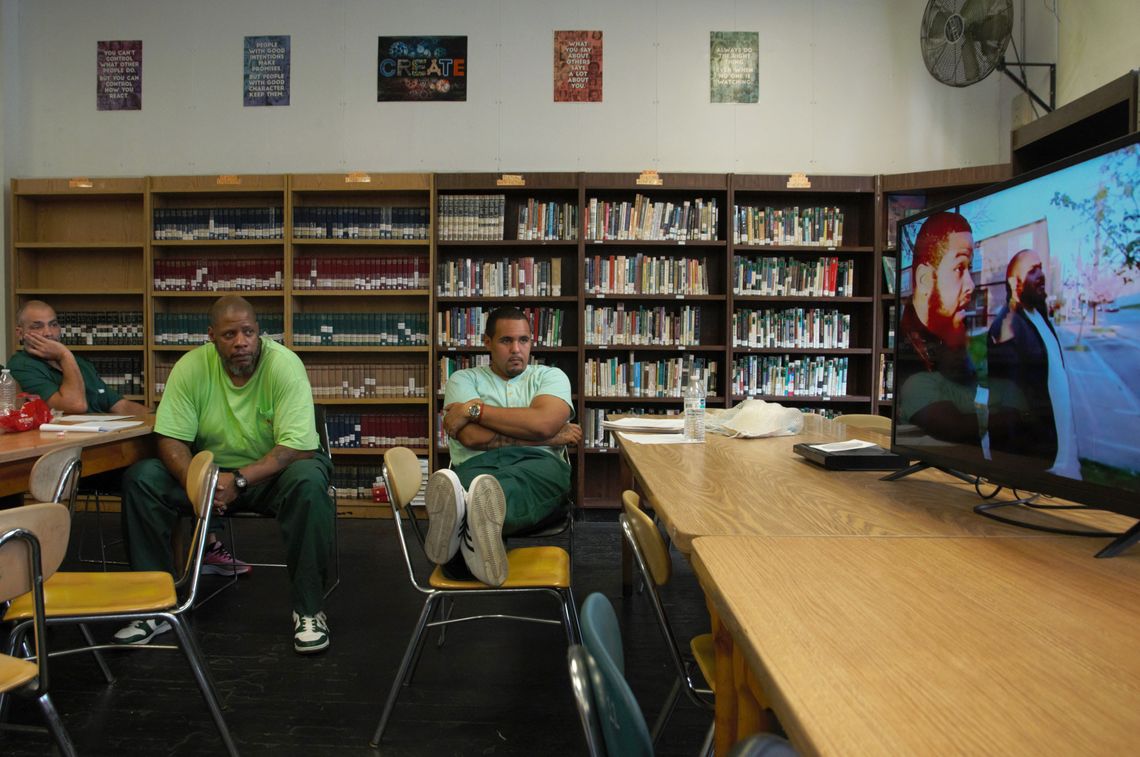 Three men wearing shades of green sit on the left while watching a film playing on a TV screen on the right. Behind the men are bookshelves filled with books.