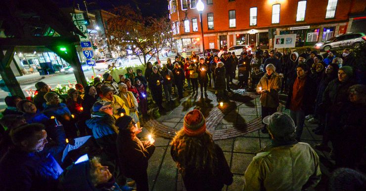 A large group of people stand in a circle on the sidewalk near an intersection, holding candles for a vigil. 