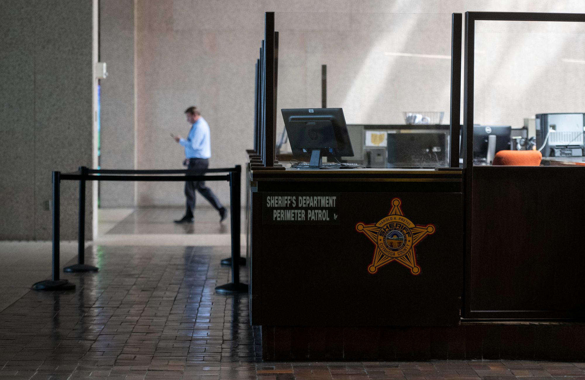 A sectioned off area inside the Cuyahoga County Justice Complex labeled “Sheriff’s Department Perimeter Patrol” has computers and chairs.  A man in a long-sleeved button-down shirt and dark-colored pants walks towards the left, behind the sectioned off area.  