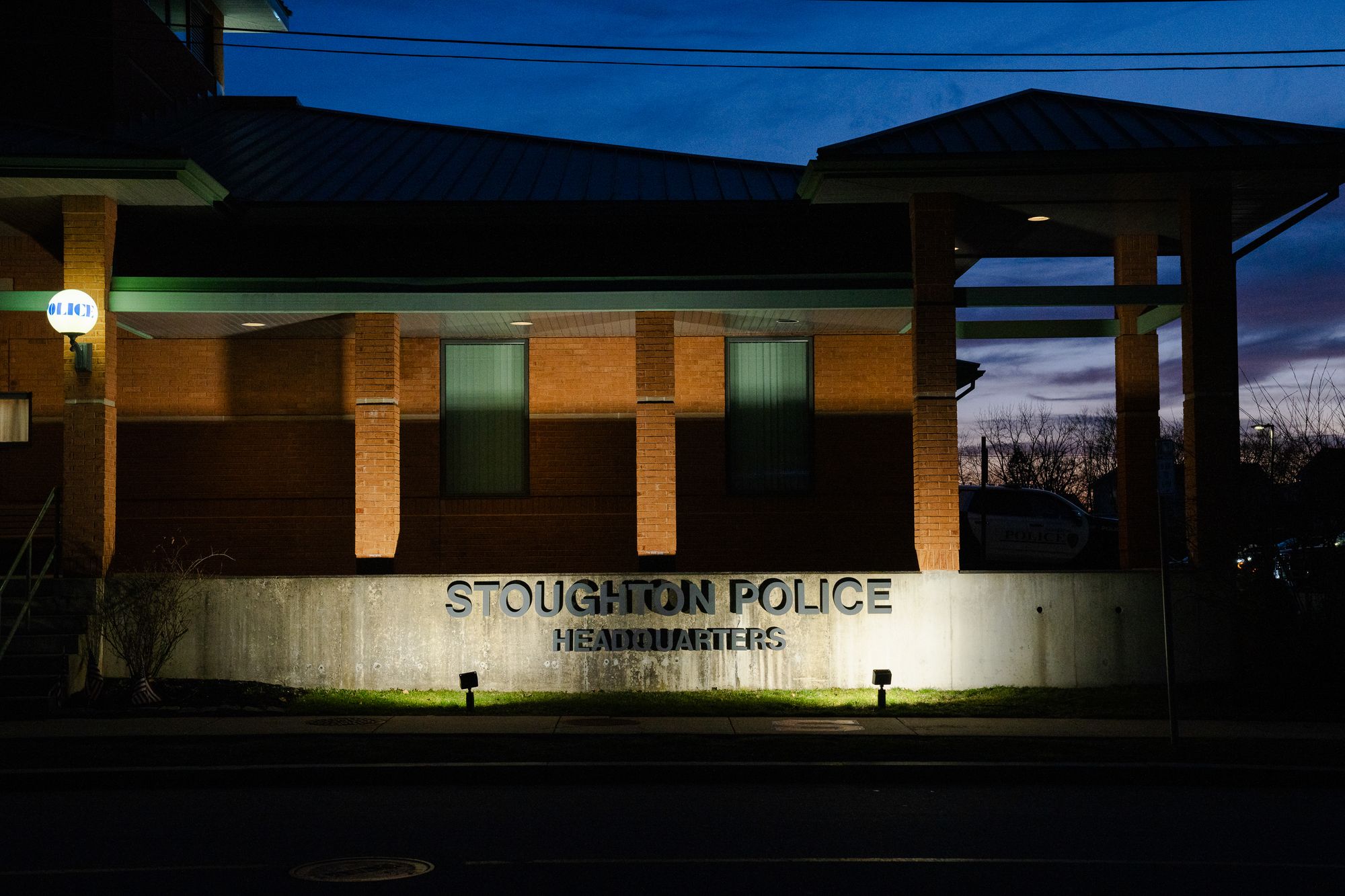 The Stoughton Police Department, a low brick building with columns, is seen at dusk, with lights showing the name of the headquarters.