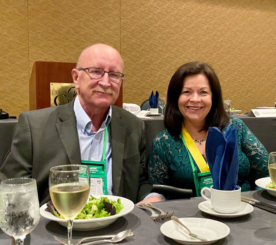 A elderly White man with a mustache, who is wearing glasses, a light blue button-down shirt, and a gray blazer, poses for a picture with his wife, a White woman with dark brown hair, who is wearing a green dress and a yellow ribbon. 
