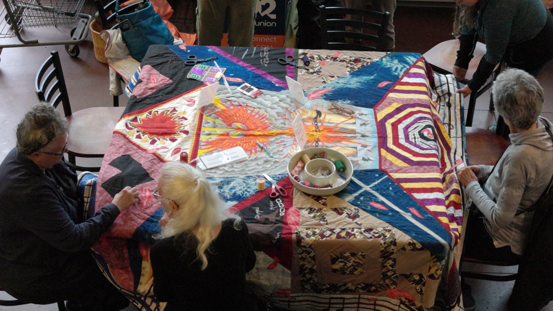 A photo shows five people sitting around a square table that has a multi-colored quilt on it.
