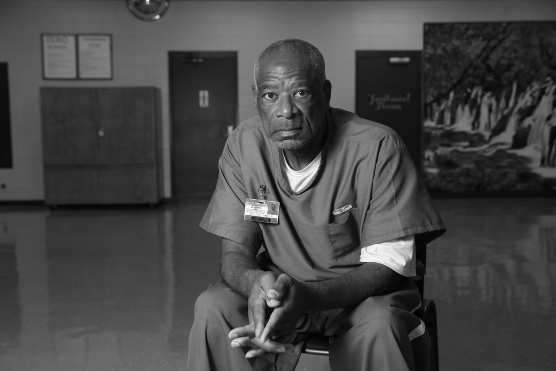 A Black man in a prison uniform sits on a chair, facing the camera for a portrait shot.