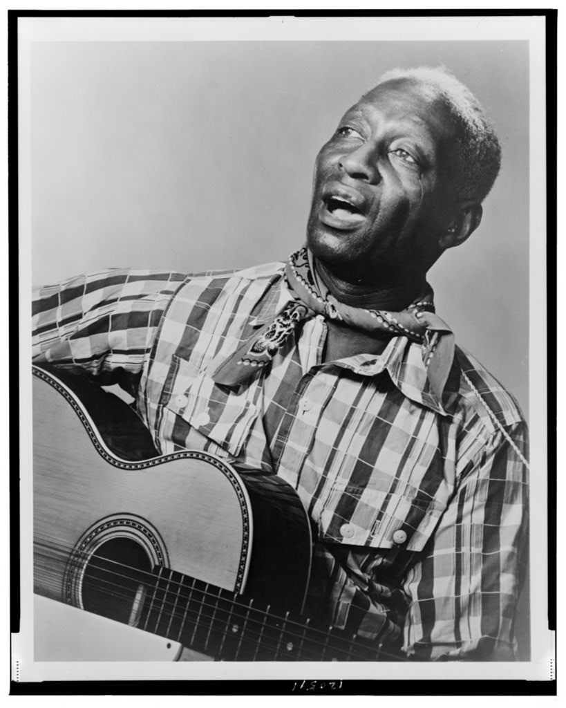 A black-and-white photo shows a Black man wearing a plaid long-sleeved shirt, holding a guitar and singing. 