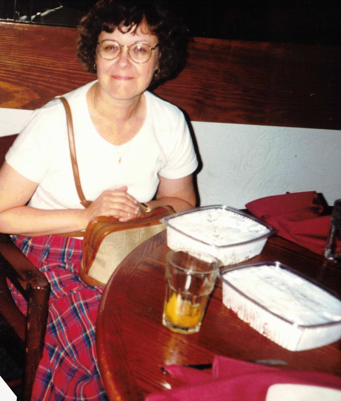A woman with brown curly hair and glasses wearing a white T-shirt and a red plaid skirt sits at a round table with baked goods.