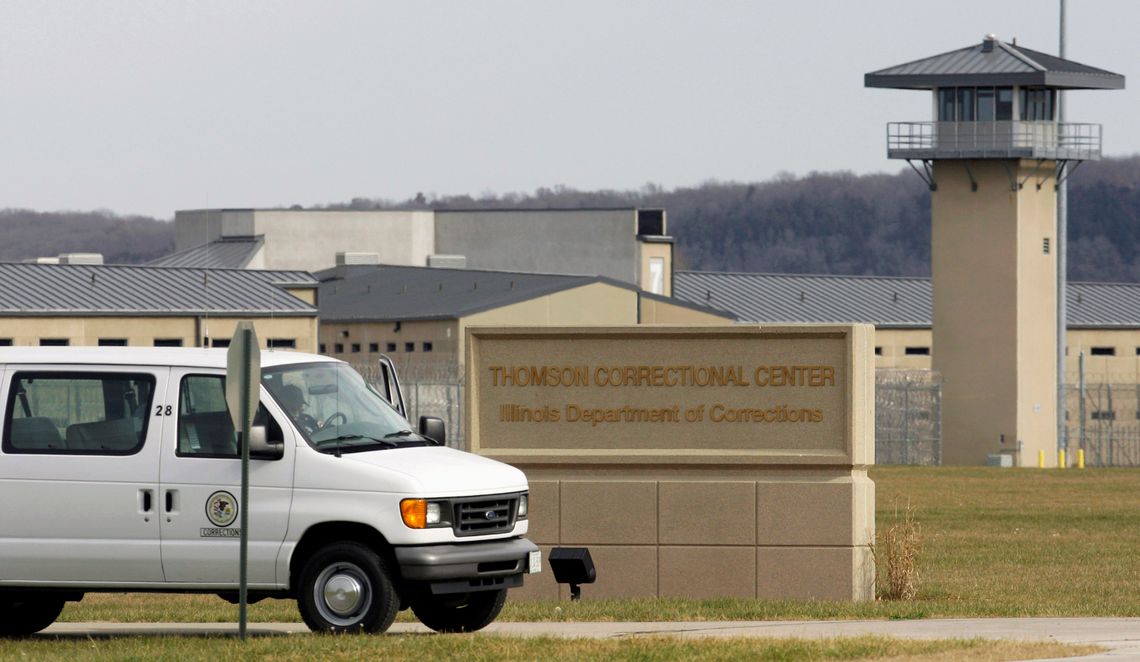 A white van drives past a beige prison building with an observation tower and a sign out front that reads “Thomson Correctional Center, Illinois Department of Corrections.” 