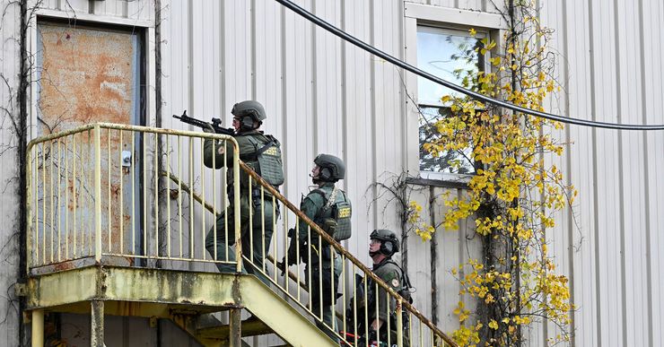 Three SWAT team members, wearing helmets, green uniforms and protective gear with the label "SHERIFF" on their backs, hold assault rifles while standing on a staircase. 
