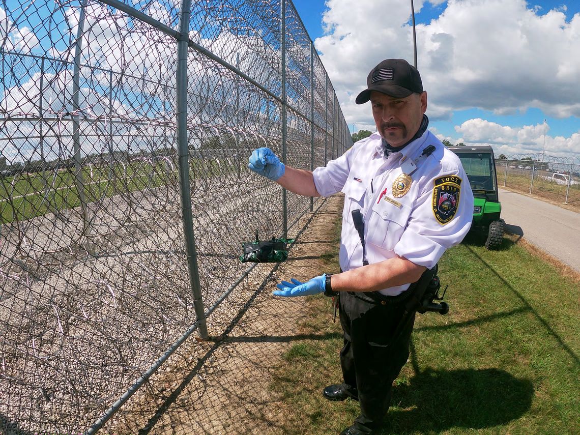 A photo shows a White man in a uniform holding a package that was brought by a drone while standing next to a prison fence. 
