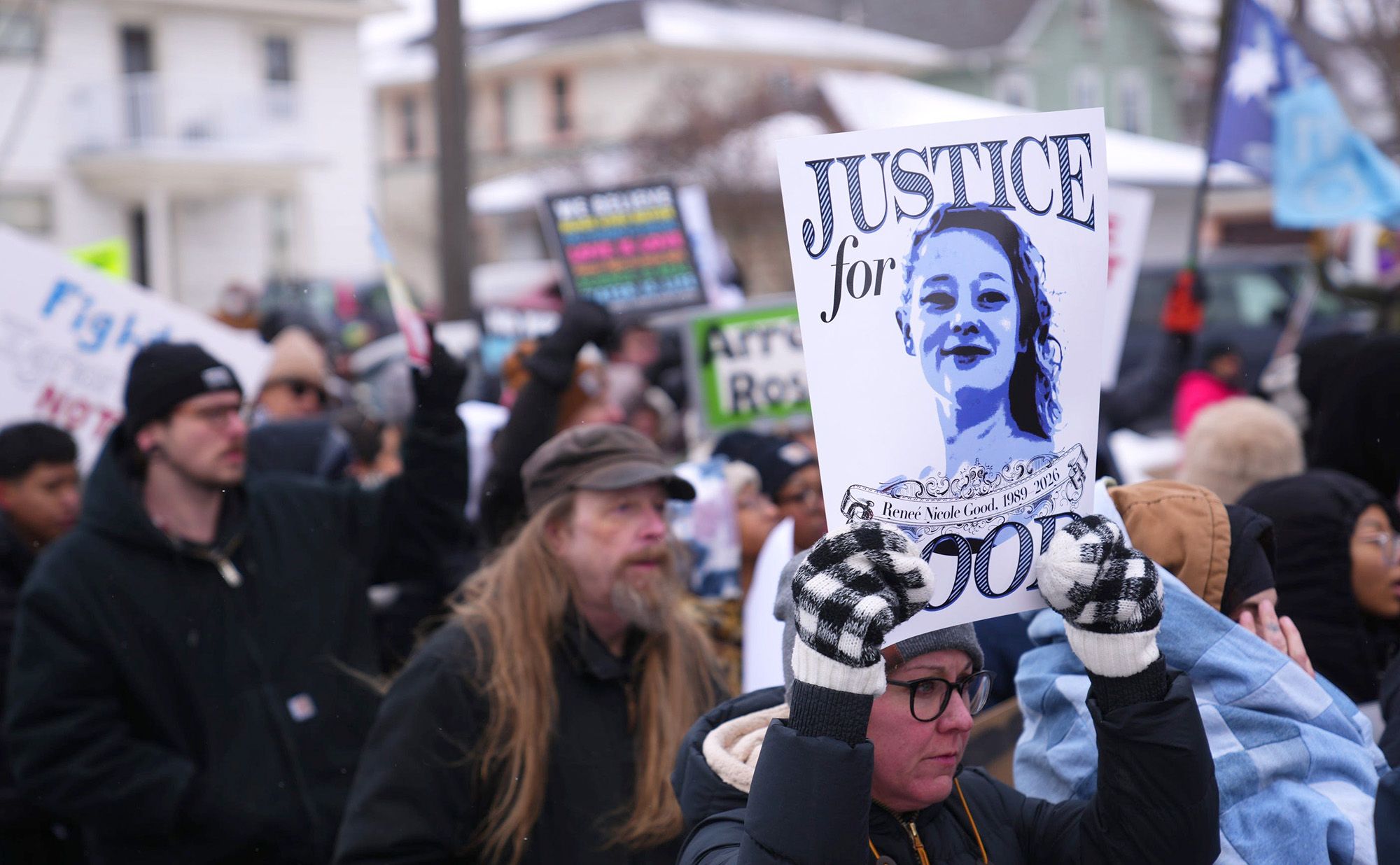 A photo of a White woman, wearing a beanie, glasses, a black puffer jacket and mittens, holds a sign that reads “Justice for Good” during a protest. Protesters holding signs stand behind her.
