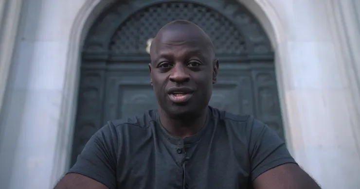 A photo shows Lawrence Bartley, a Black man in a dark gray T-shirt, speaking outside in front of a doorway.  