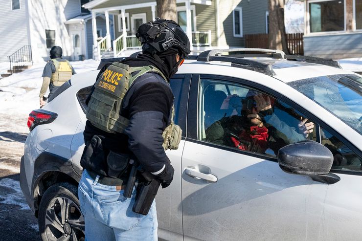 A photo shows a U.S. Border Patrol agent, wearing head gear and a vest, with their hands on their hips as they look at a passenger inside a car. The passenger is holding up a phone, recording the agent. 