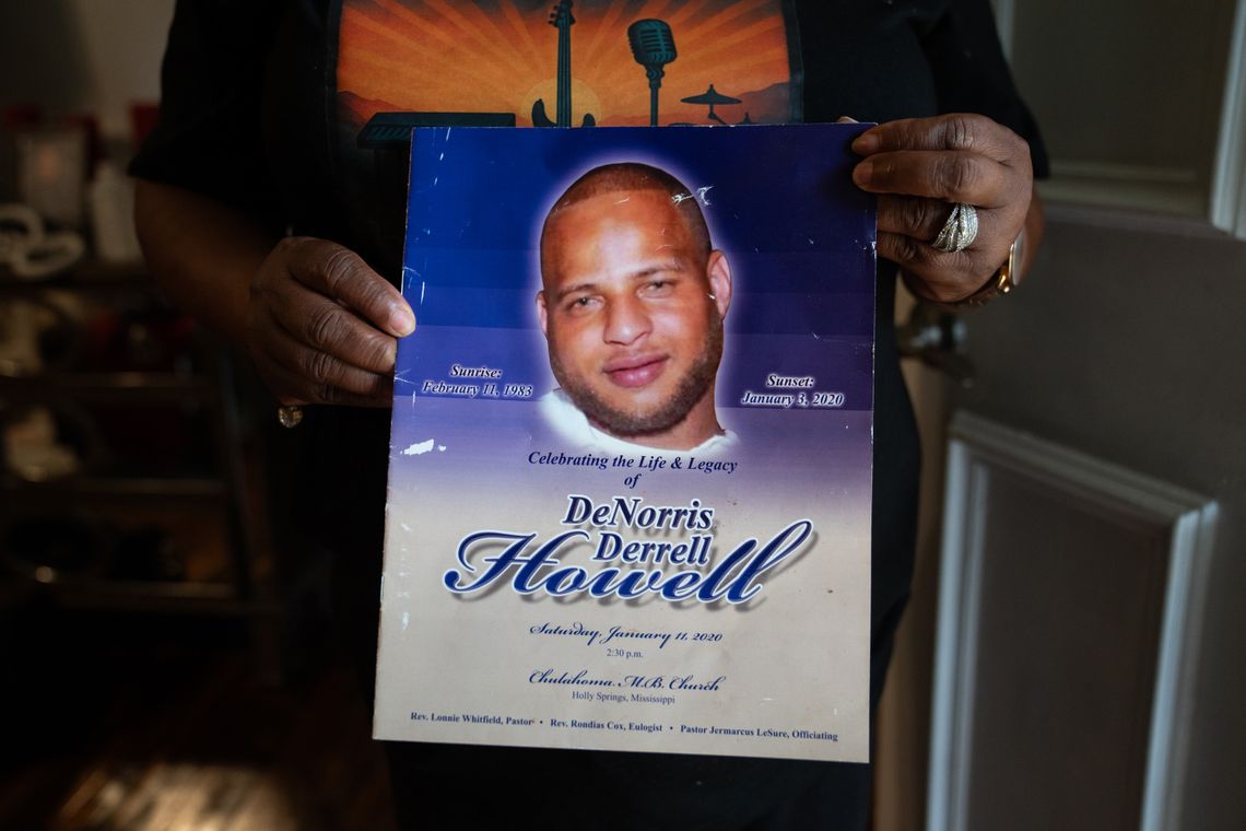A photo shows a close-up of a Black woman’s hands holding a funeral program for Denorris Derrell Howell. The funeral program features a cutout portrait of a bald, Black man with a goatee on a blue background. 