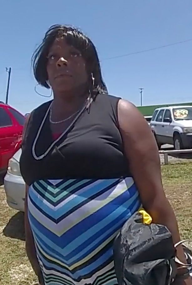 A Black woman, wearing a black and blue patterned dress, is standing while holding a black bag.