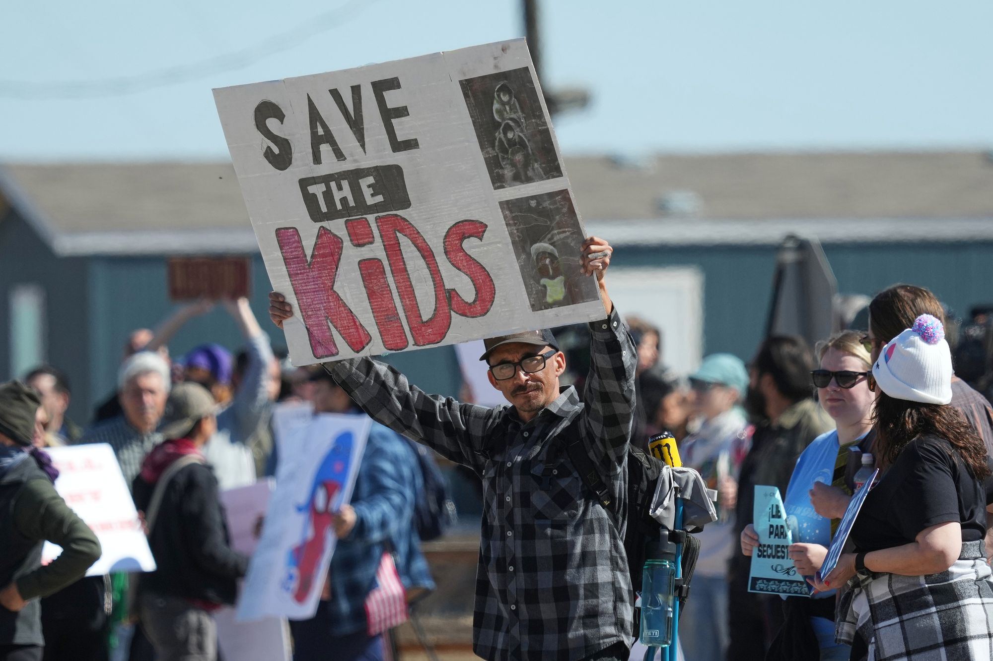 A photo shows a man with medium-toned skin, wearing a baseball cap and a plaid shirt, holding a sign that says, “Save the Kids.” He stands among other protesters who are holding signs. A low bluish building with a gray roof is behind them.  