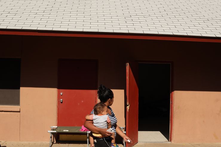 A photo shows a woman with medium skin tone and black hair with her head turned toward an open red door. She holds an infant in one arm.