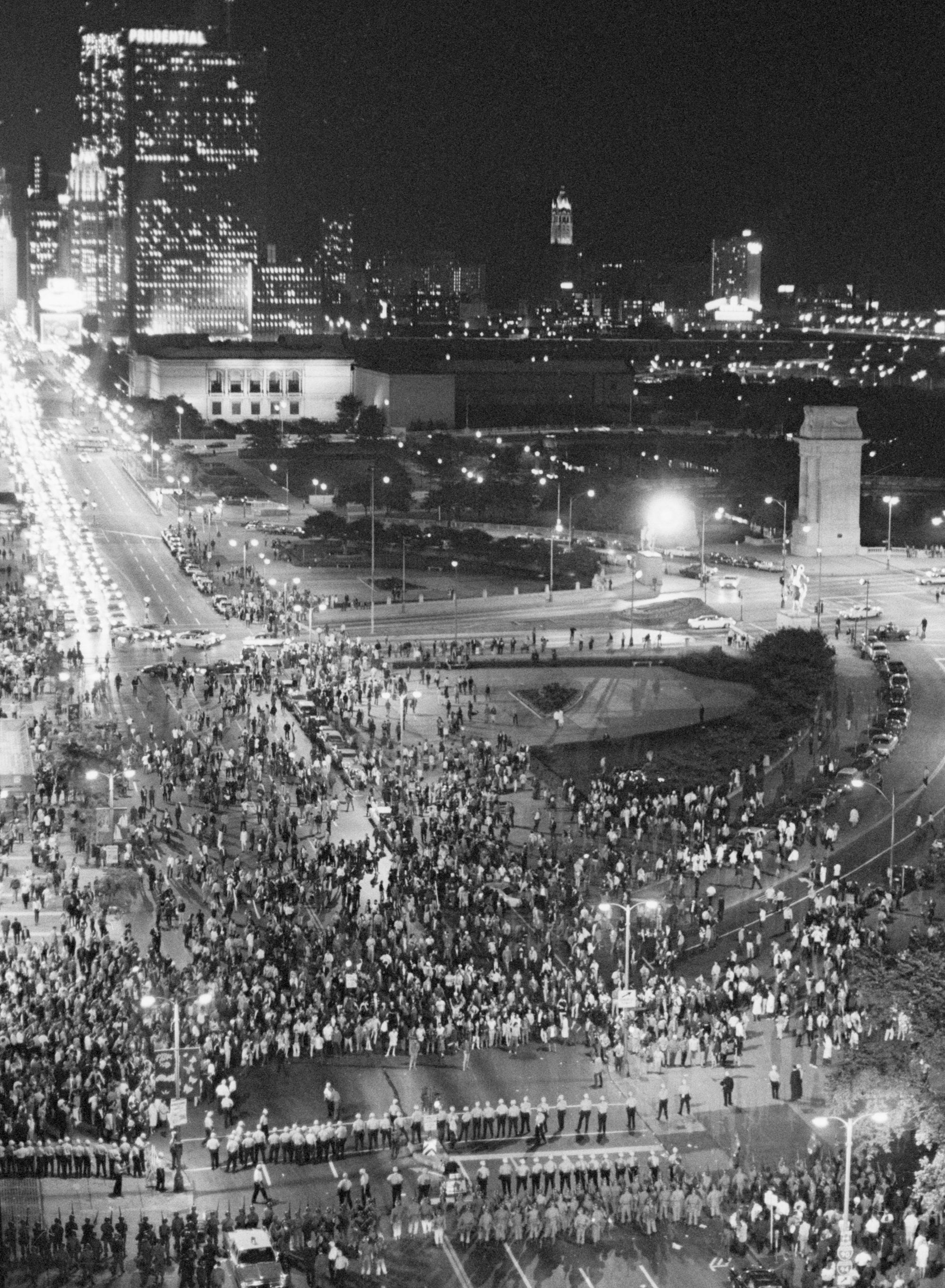 A black-and-white photo shows an aerial night view of a broad street next to a park, with tall buildings in the background. Thousands of demonstrators are marching in the street and park as police and National Guardsmen confront them.