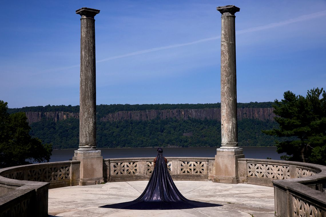 Khayr Muhammad stands at an overlook in the gardens, wearing a dark purple cape that spreads out behind him. 