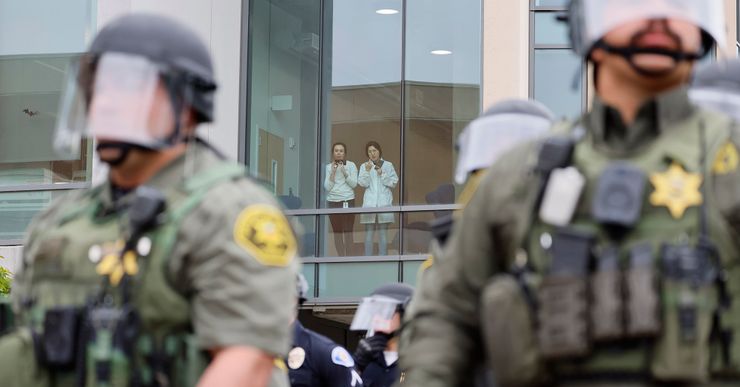 Two university members, wearing white tops, jeans and badges, look out a window and record videos from their phones. In the foreground, police wearing riot gear form a line in front of a building. 