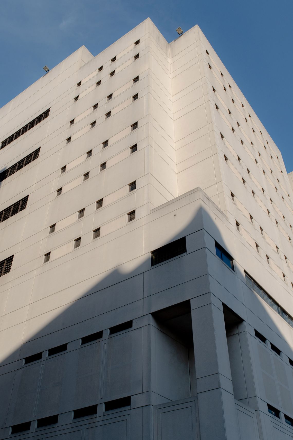A photo, taken from a low angle, shows the outside of a light gray tower with small, square windows. Blue sky is visible above the building. 
