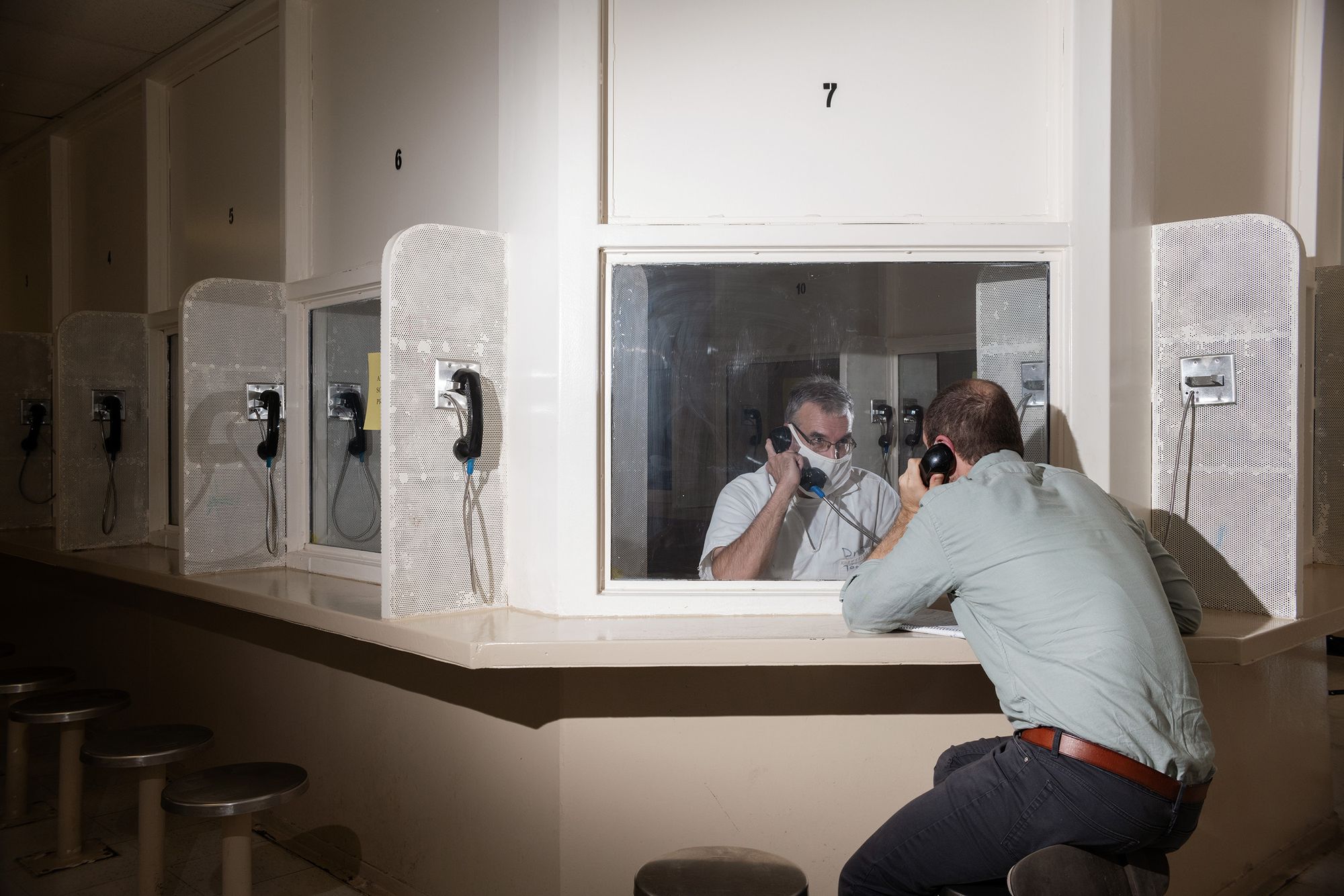A photo shows Larry Driskill, a White man wearing a white uniform and a facemask, talking on a phone from behind glass to Maurice Chammah, a White man wearing a pale green shirt and dark pants, who is seated across from Driskill and also holding a phone, in a prison. 