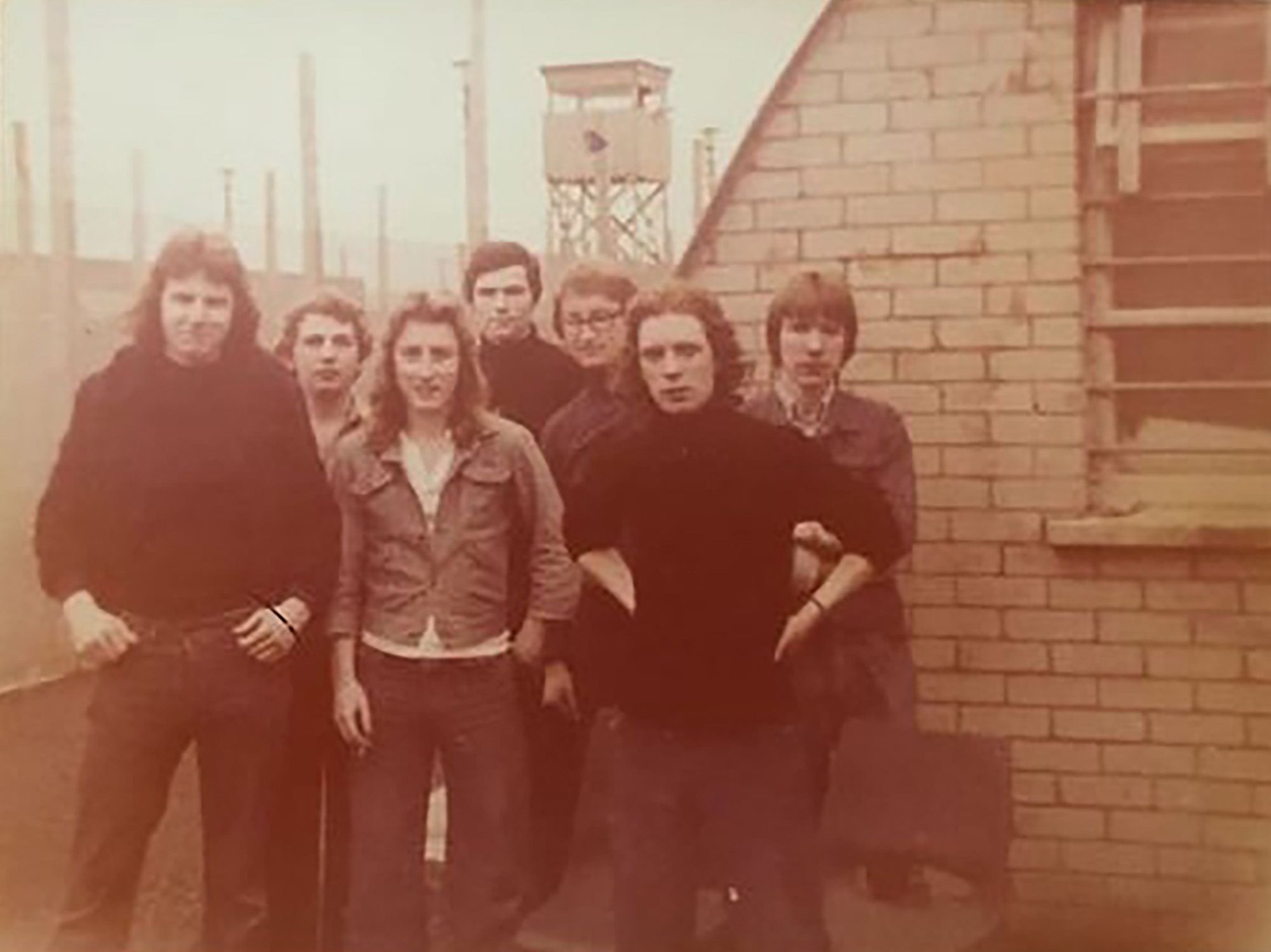A sepia-toned photo shows a group of young adults, wearing black and gray clothes, standing in front of a brick building. There are several poles and a watchtower in the background. 