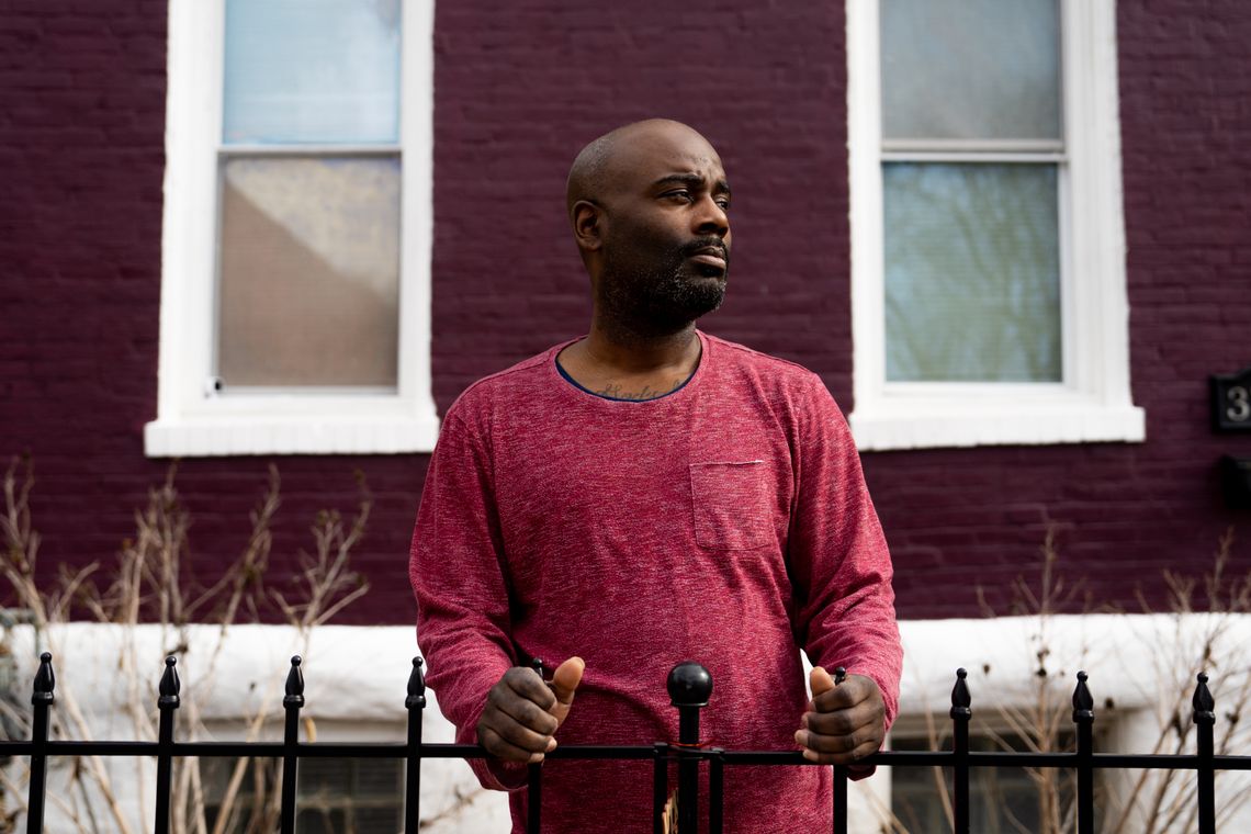 A Black man with a red shirt looks away while standing outside his brick house painted purple and white.
