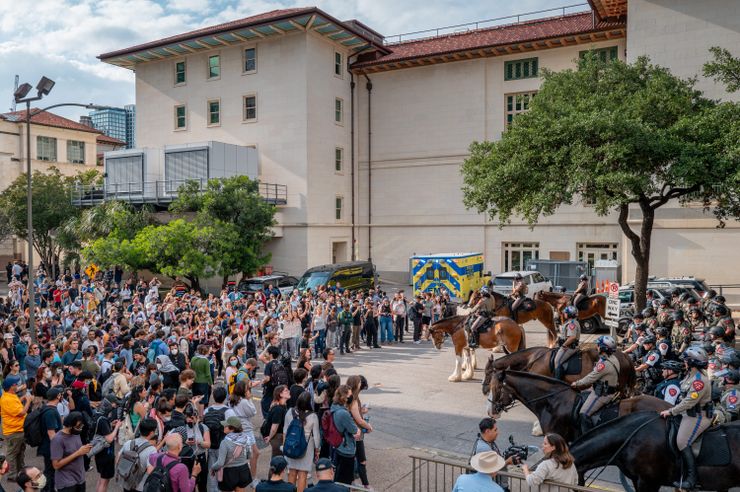 A photograph shows a large group of students facing toward police wearing riot gear on a college campus. Some police officers are mounted on horses. 