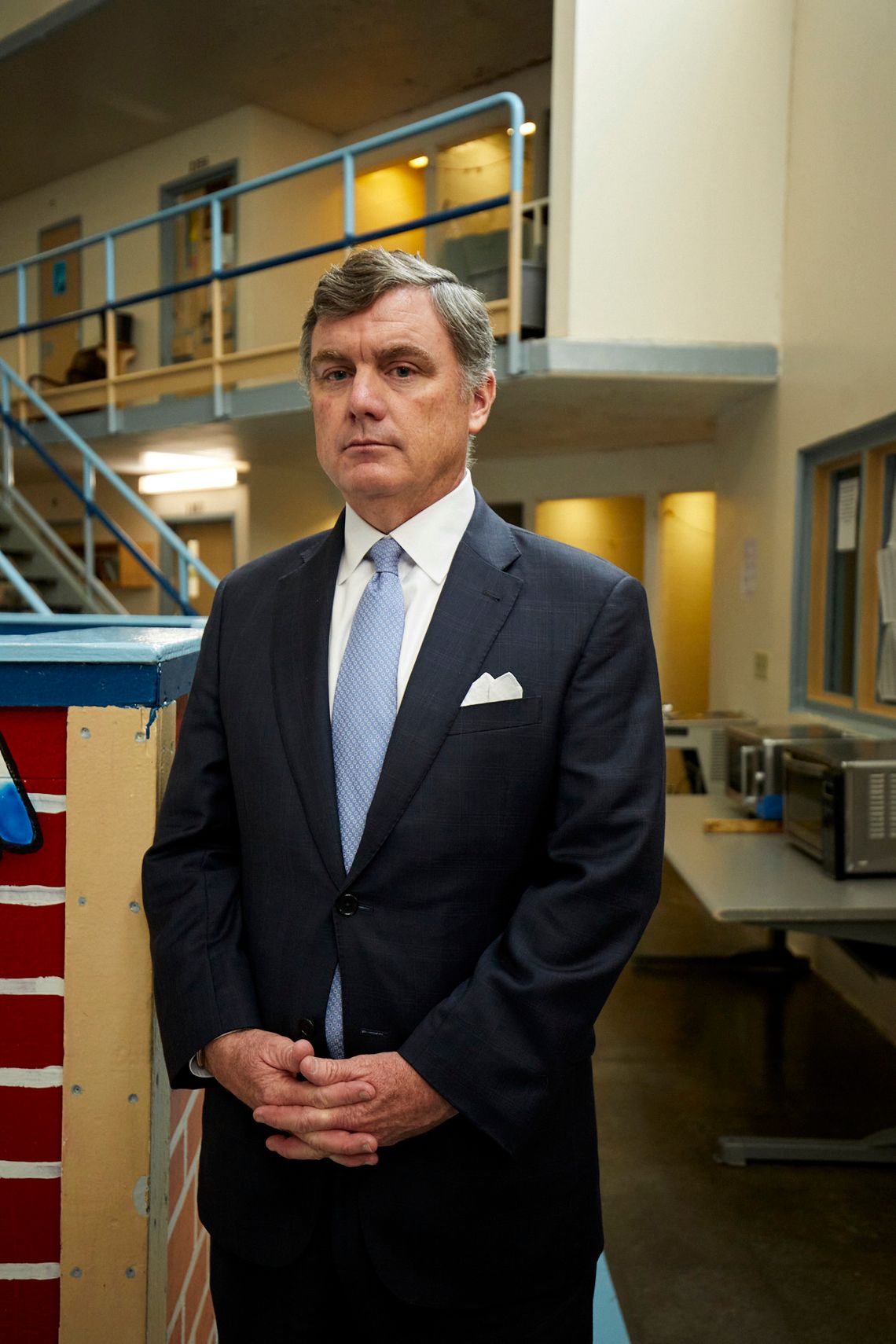 A vertical photo of Bryan Stirling, a White man with short gray hair wearing a black suit and light blue tie, stands in the Lee Correctional Institution.