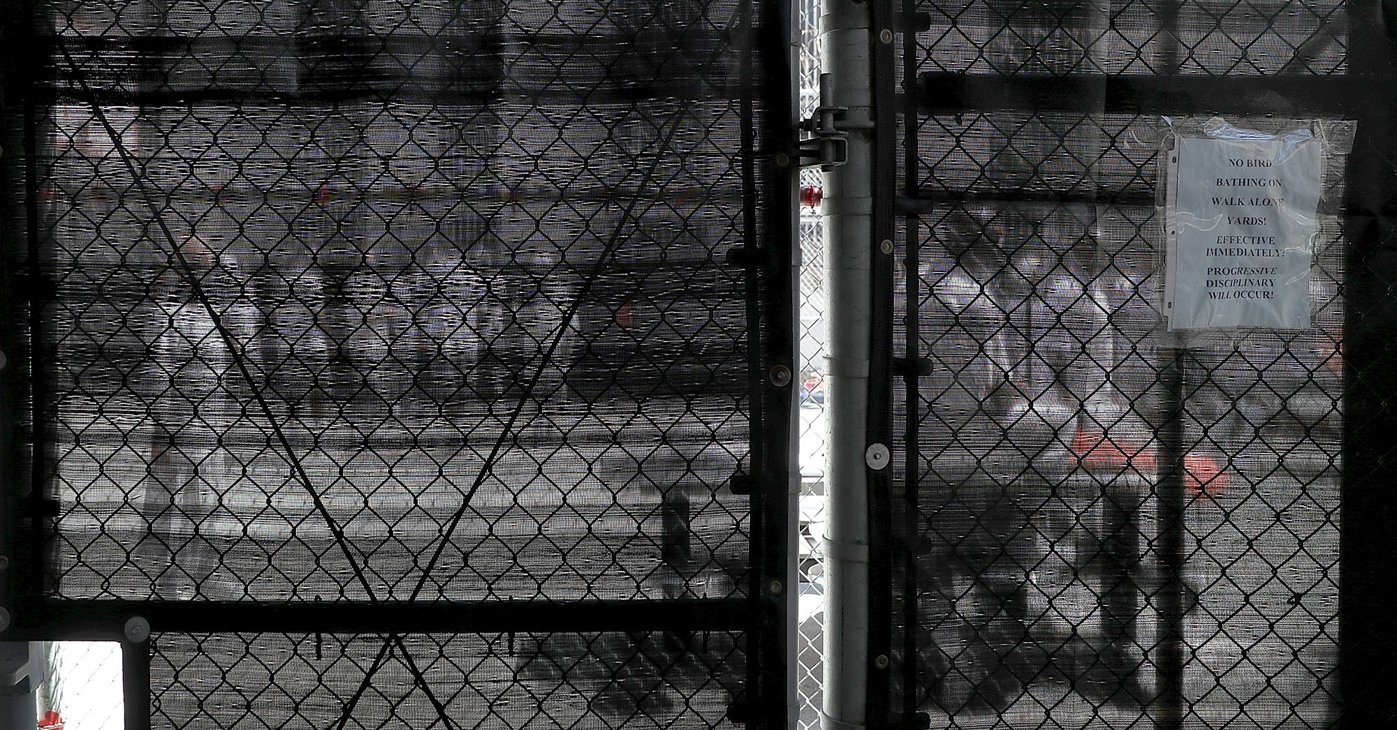 A photo shows incarcerated people seen through a fence covered in black mesh, standing in a yard outside.