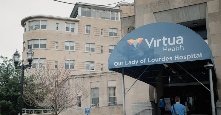 A person wearing a blue uniform and black messenger bag walks toward the entrance of a hospital with a blue sign that reads, "Virtua Health Our Lady of Lourdes Hospital."