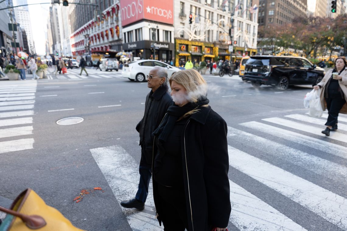 A photo shows a White woman, Sandra Hafraoui, with medium-length blonde hair, crossing the street in New York City. Vape smoke is coming out of her mouth. Cars and shops, including a Macy’s department store, are visible behind her. 