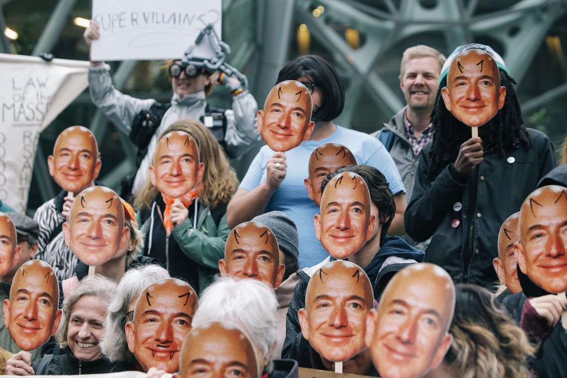 A photograph shows a group of people holding up posters and masks of Amazon founder Jeff Bezos, a White, bald man, with drawn-on devil horns in front of their faces. 