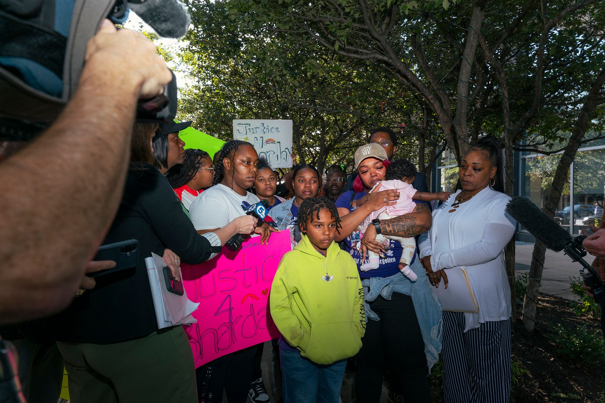 A group of people are gathered at an outdoor press conference. Two hold signs reading "Justice for Sharday." One woman with tears streaming down her face holds a baby. News video cameras are in the foreground.