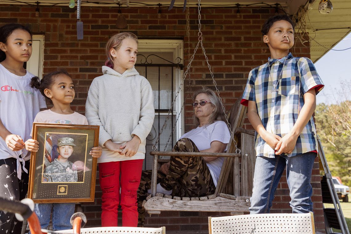 A group of four multiracial children stand on a porch of their grandparent’s home. One of them holds a framed portrait of their mother in a U.S. Army Reserve uniform. In the background, a White elderly woman, wearing glasses, white T-shirt and leggings, is sitting on a bench. 
