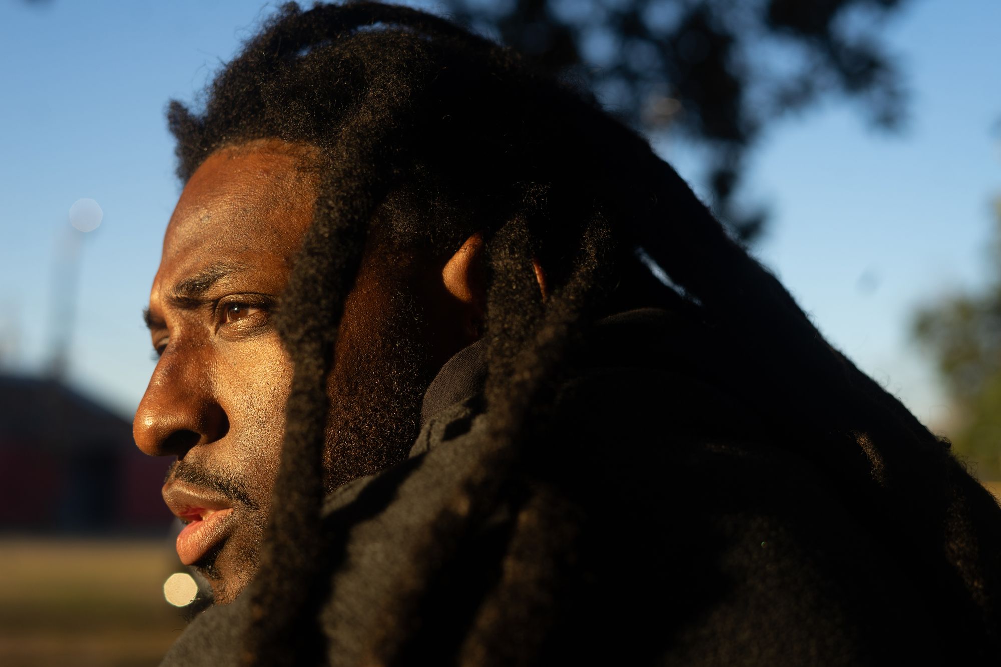 A Black man with dreads sits outside with the sunlight on his face.