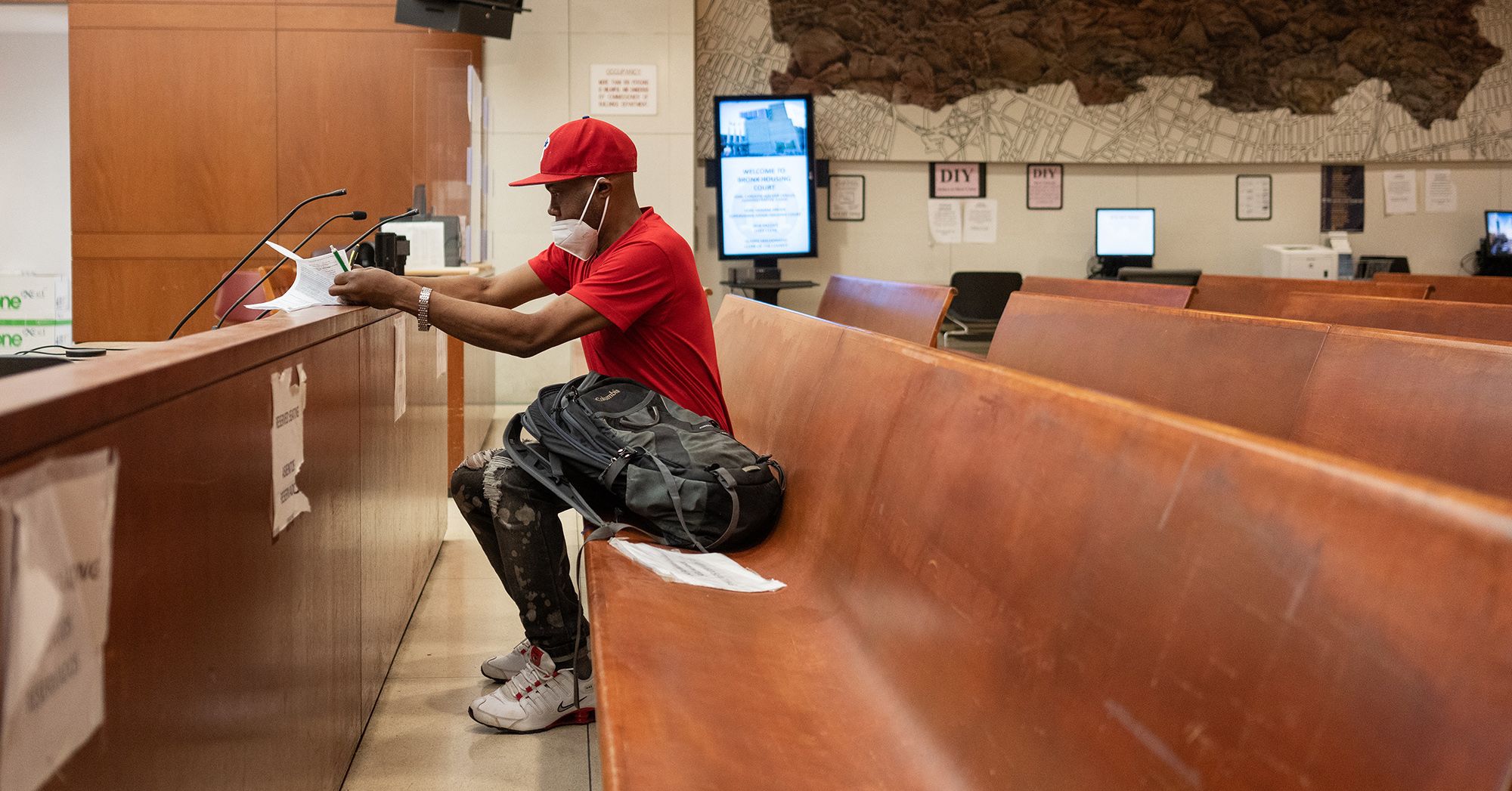 A Black man, wearing a KN95 mask, red shirt, red cap and camo pants, reviews a document while sitting in a courtroom. 


