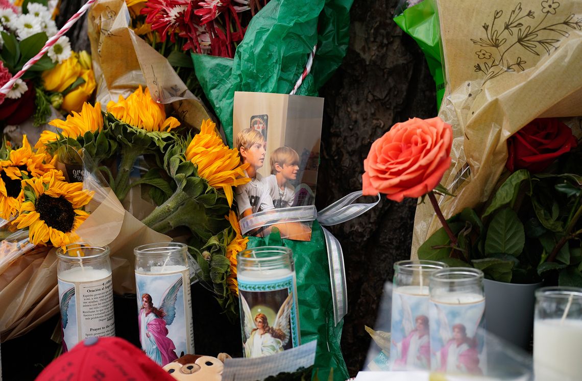 A memorial is filled with candles, a picture of two young boys, baseballs, and flowers at the base of a tree. 