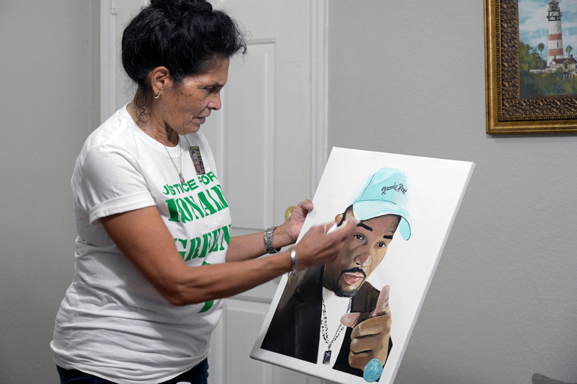 A woman with medium-toned skin and dark hair holds a painting of her son, Ronald Green, a Black man wearing a blue baseball cap.  