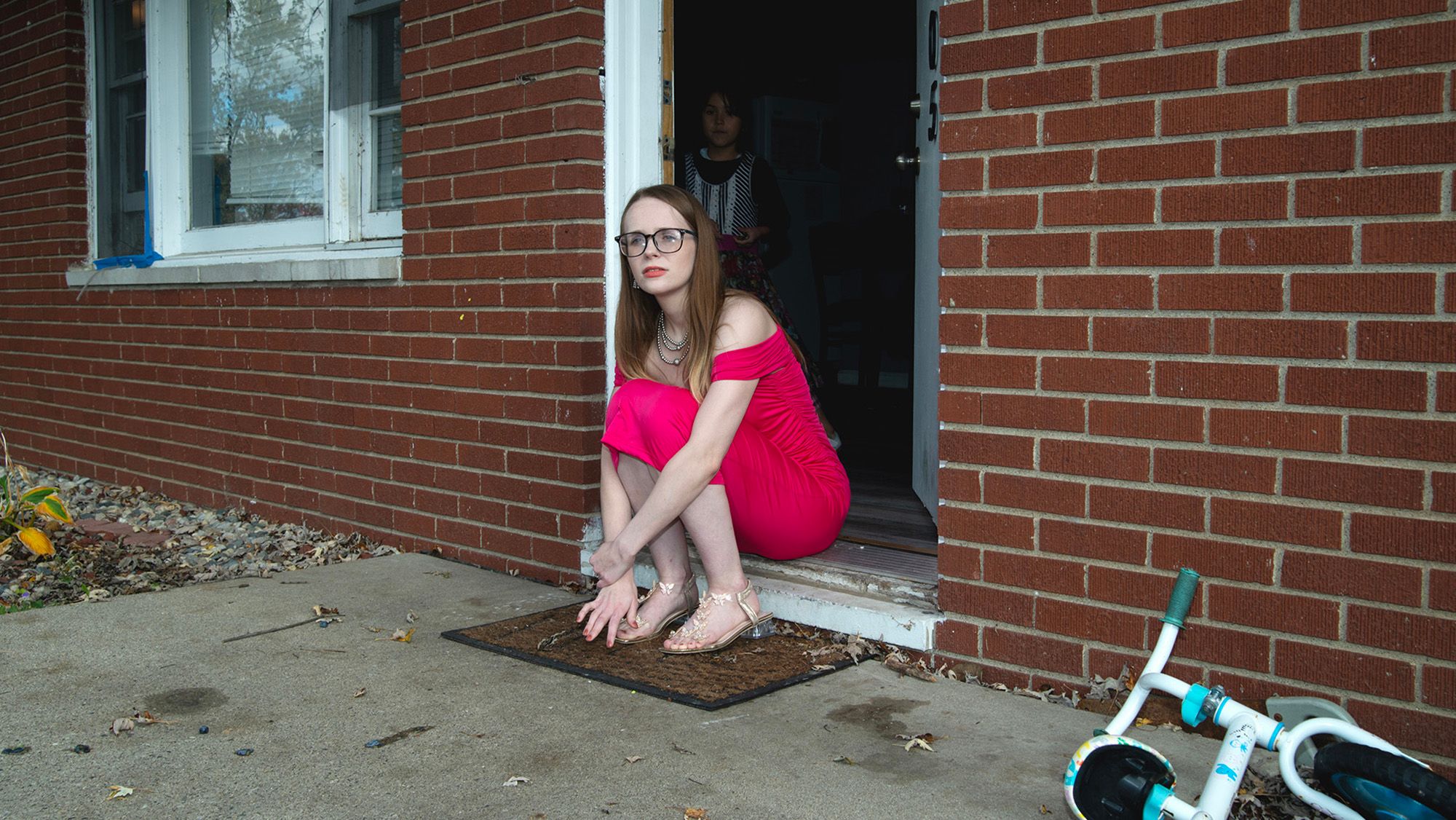 A White woman, wearing glasses and a red dress, sits at the front door of her house. One of her children stands behind her inside. A children's bike is stationed on the right side of the door.