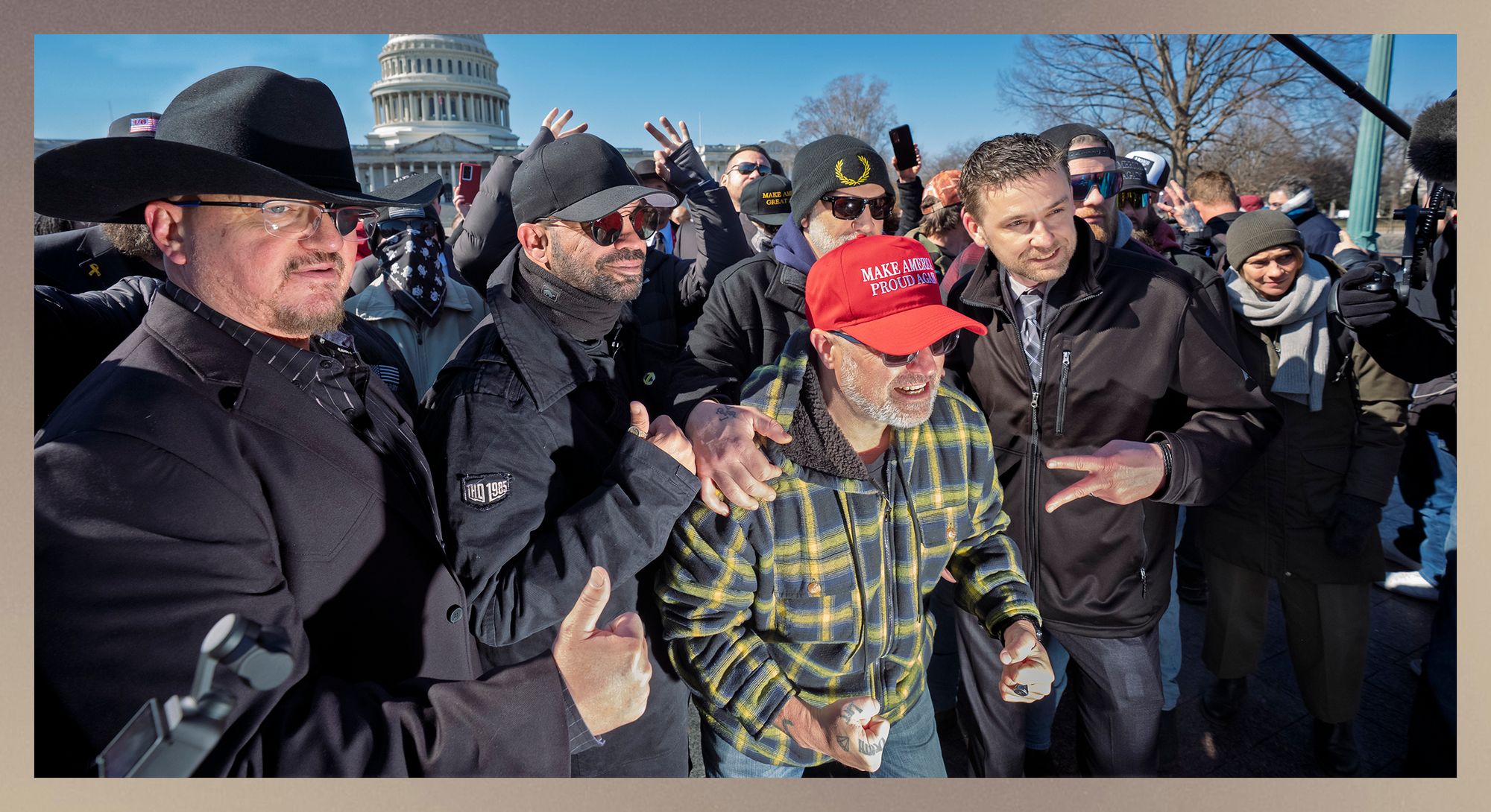 A group of men, including from left, Stewart Rhodes, a man with light-toned skin, wearing a black hat and glasses with a patch over his left eye, and Enrique Tarrio, a man with medium-toned skin, wearing sunglasses and a black baseball cap, stand outside in Washington, D.C. Both have their thumbs up. 
