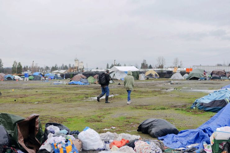 Two people walk through an encampment for unhoused people, which is on a field with some patches of grass and dirt.  Tents are visible both in the distance and in the foreground, and they stand beside the residents' belongings.    