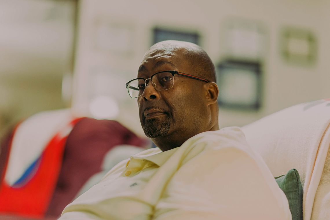 A photo of Gerald Laird, a bald, Black man with a goatee and a cream-colored shirt, sitting on a couch in his living room. Framed certificates are out of focus behind the man. 