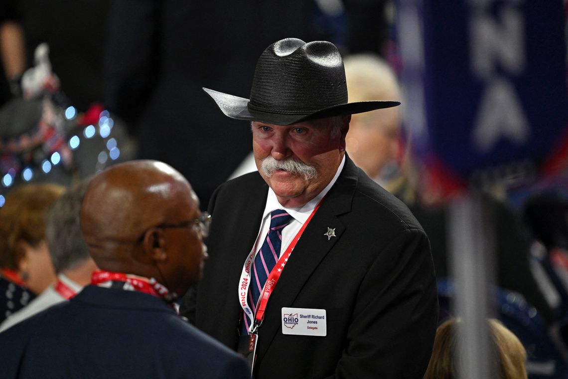 A photo shows a White man with a moustache, wearing a black cowboy hat and a suit, standing in a group of people. 