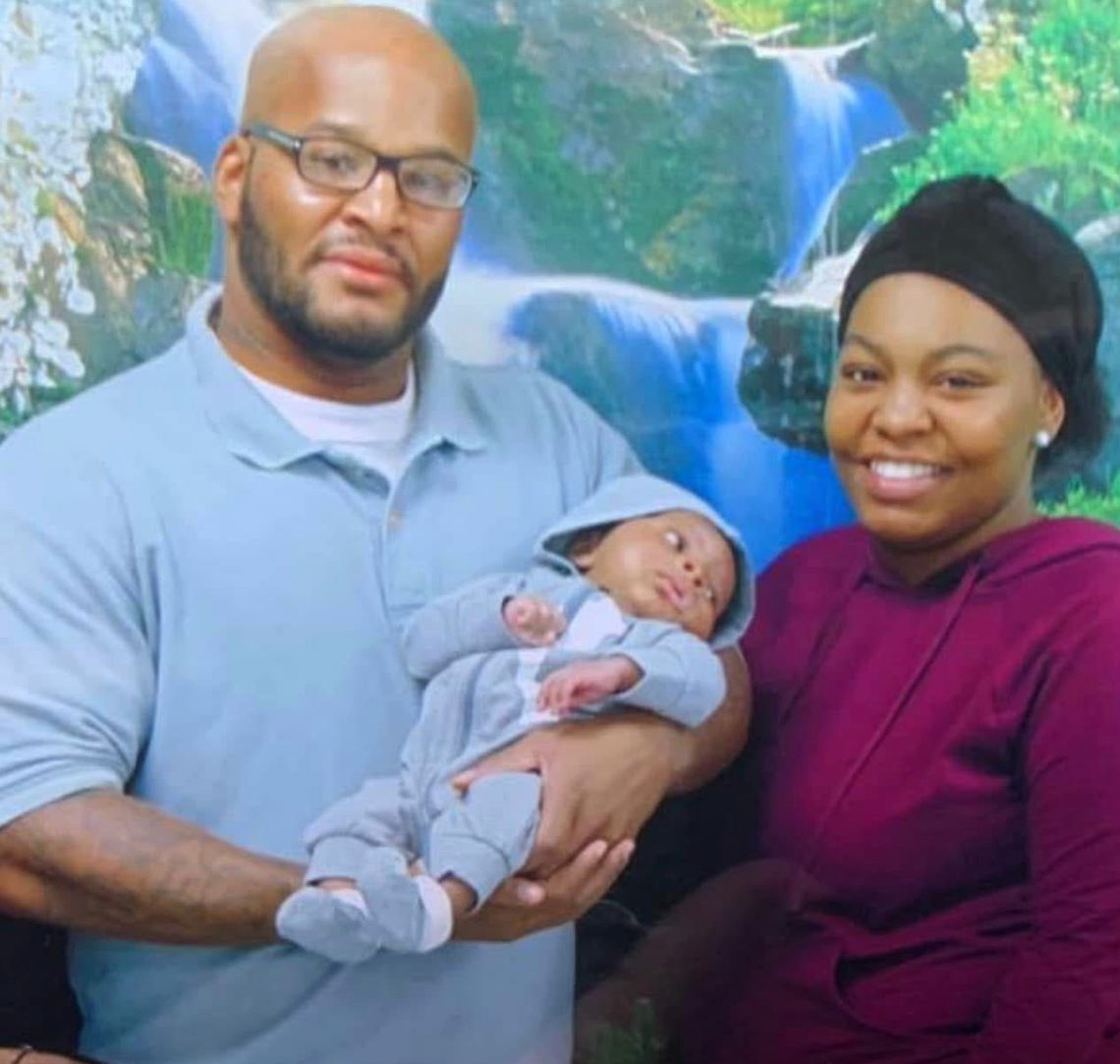 A Black woman in a black headwrap and a magenta sweater smiles for a picture as she stands next to her father, who is wearing a gray prison uniform. Her father is holding a baby. 