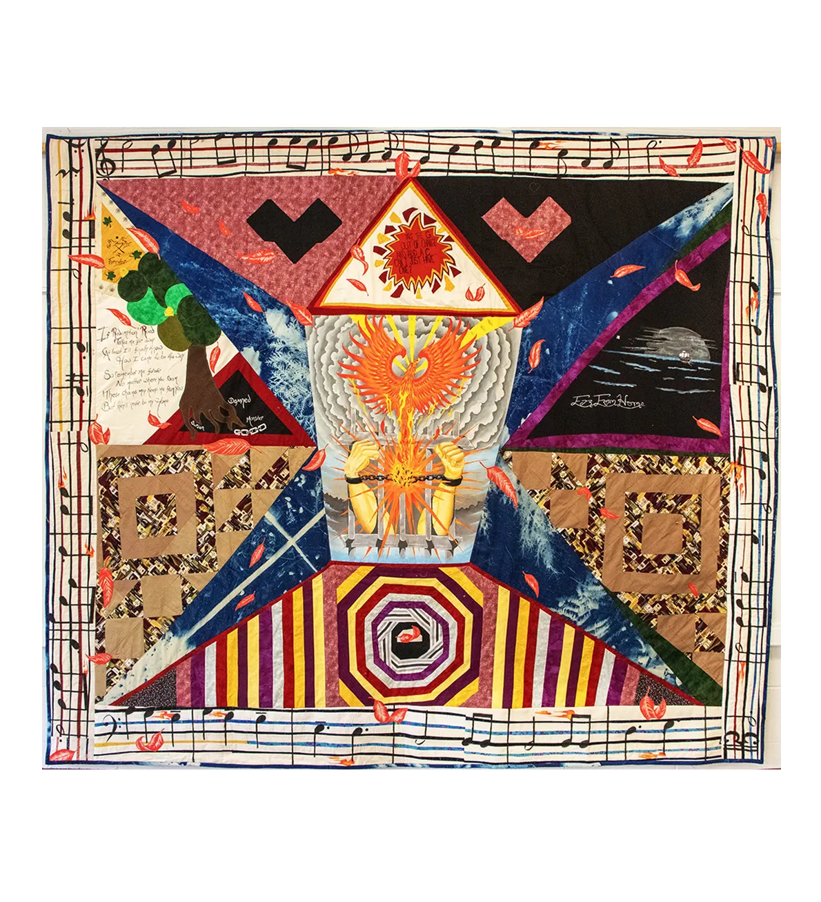 A photo of a quilt shows, at center, two arms with raised fists in handcuffs reaching out from behind prison bars. There is a phoenix rising from the center of the handcuffs, where there is a starburst. Multi-colored pieces surround the center, and the edges of the quilt are lined with a pattern of musical notes on a staff.